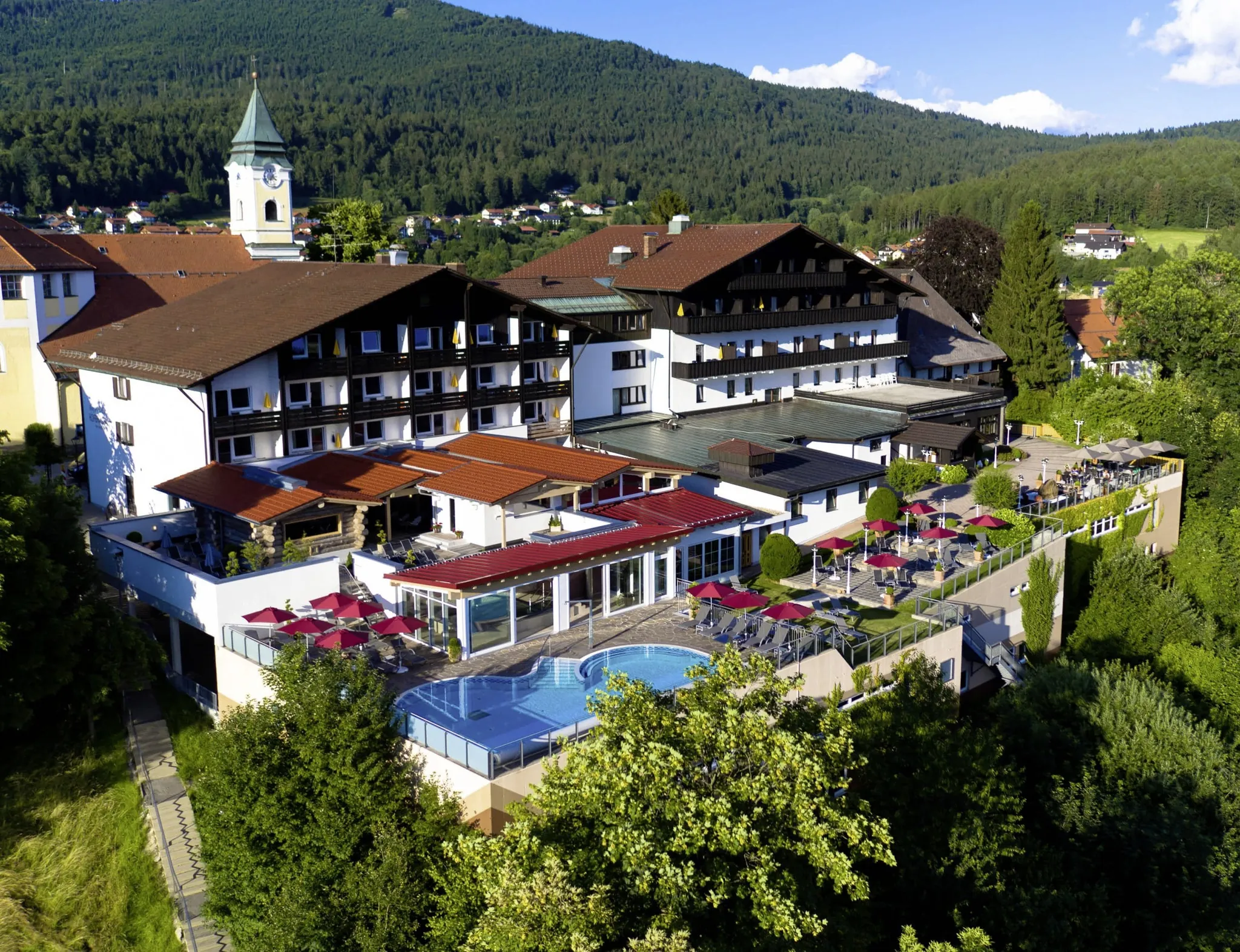Außenansicht des Wellnesshotel Hofbräuhaus mit roten Sonnenschirmen und Außenpool, umgeben von grünen Bäumen und Bergen im Hintergrund