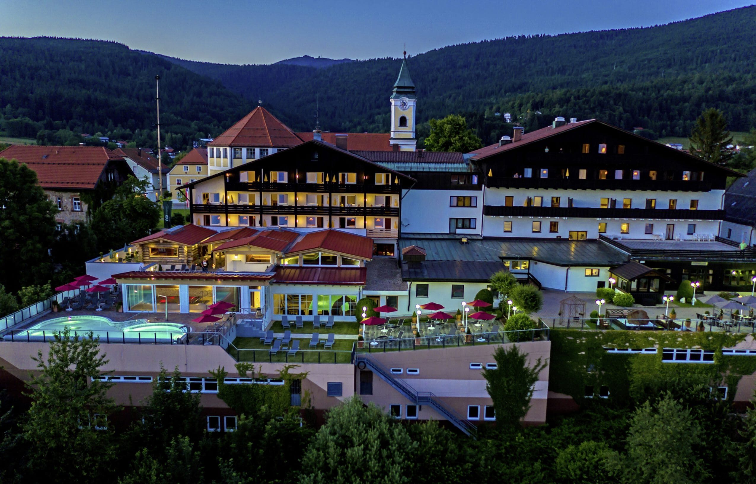 Beleuchtete Hotelfassade des Wellnesshotel Hofbräuhaus mit Außenpool und Liegestühlen vor bewaldeten Hügeln bei Abenddämmerung