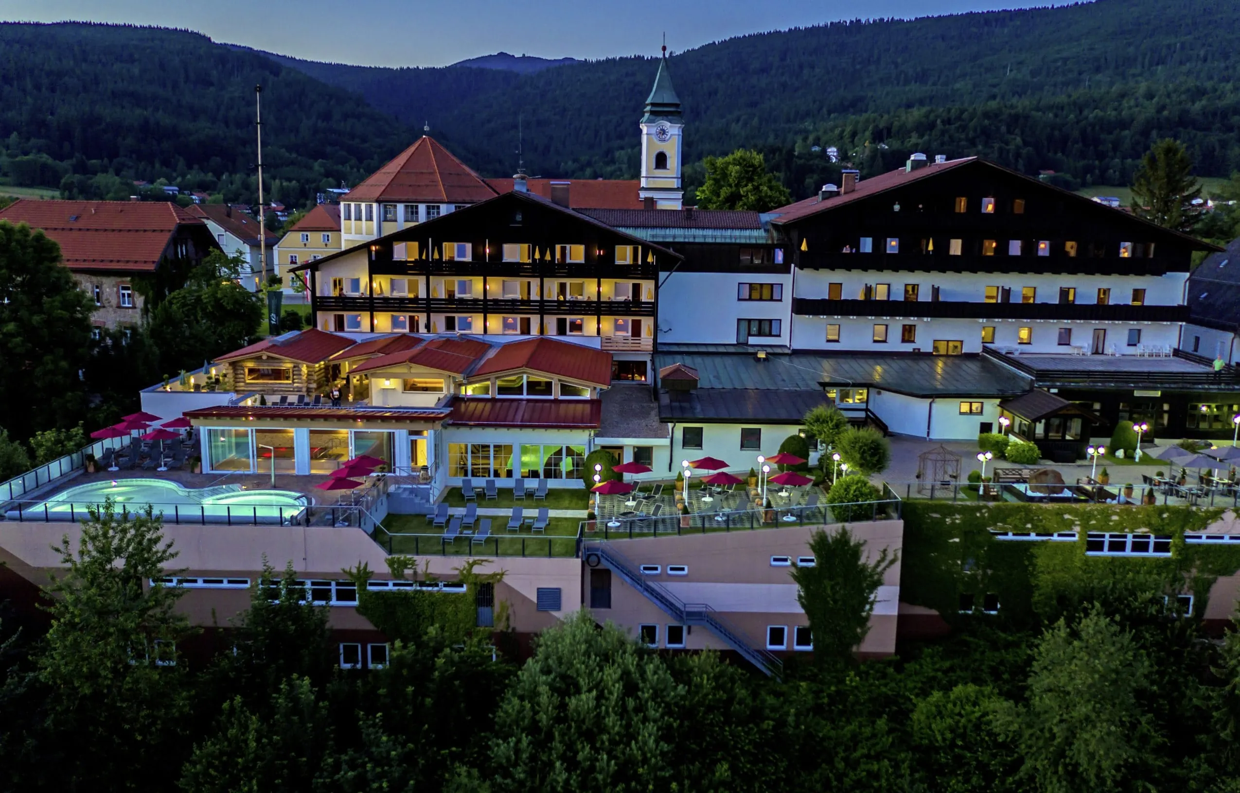 Beleuchtete Hotelfassade des Wellnesshotel Hofbräuhaus mit Außenpool und Liegestühlen vor bewaldeten Hügeln bei Abenddämmerung
