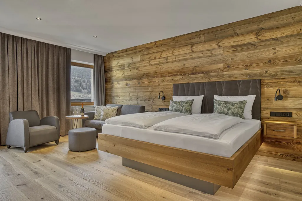 Modernes Hotelzimmer des Wellnesshotel Hofbräuhaus mit Doppelbett, Holzwand, grauen Vorhängen, Sessel, Sofa und kleinem Tisch mit Flasche und Gläsern