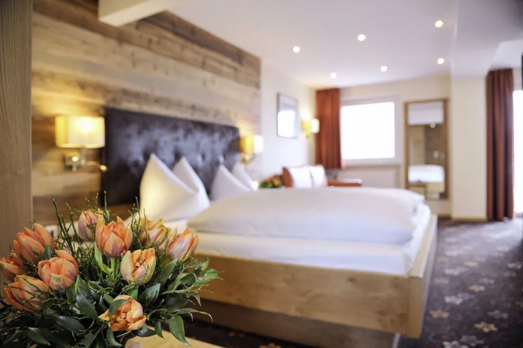 Hotelzimmer des Wellnesshotel Randsbergerhof mit großem Bett, Holzwand mit Kopfteil, oranger Blumendekoration und Fenster mit roten Vorhängen