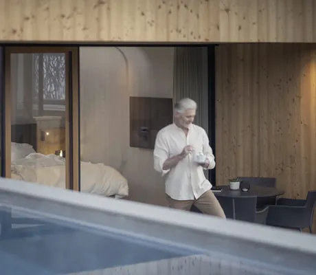 Mann mit Kaffeetasse in der Hand steht auf der Terrasse eines Hotelzimmers des Hotel Winkler mit Blick auf das Doppelbett