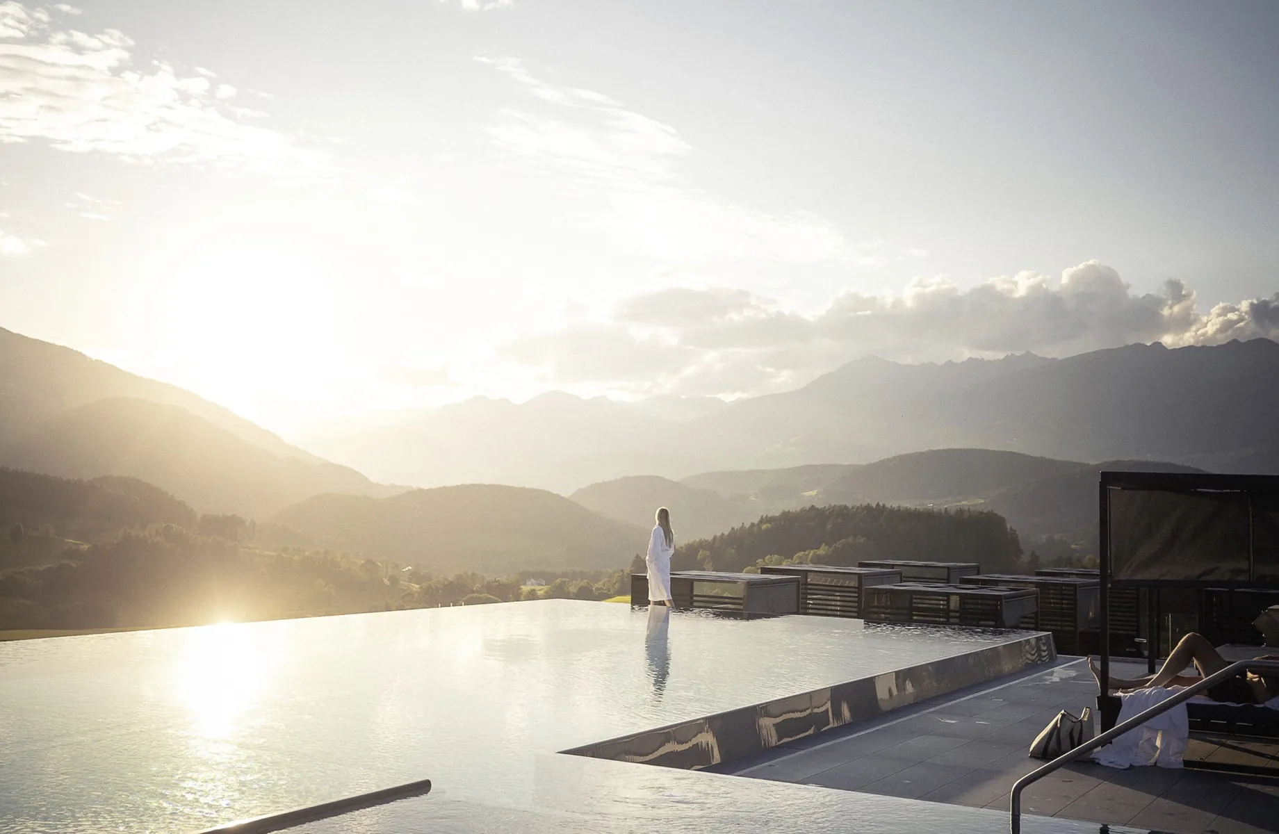 Frau in weißem Bademantel steht am Skypool des Hotel Winkler mit Blick auf die Berge bei Sonnenaufgang