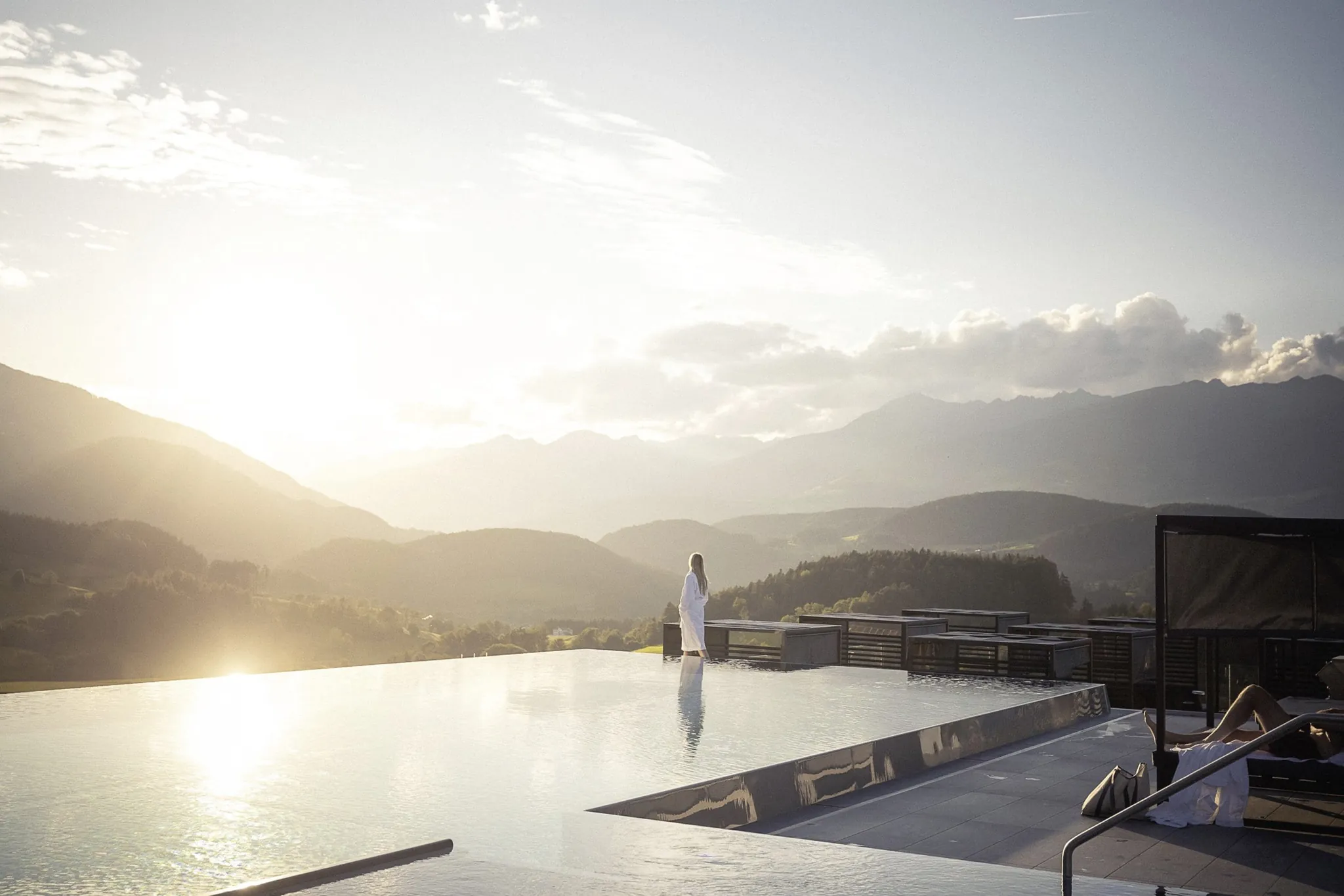 Frau in weißem Bademantel steht am Skypool des Hotel Winkler mit Blick auf die Berge bei Sonnenaufgang