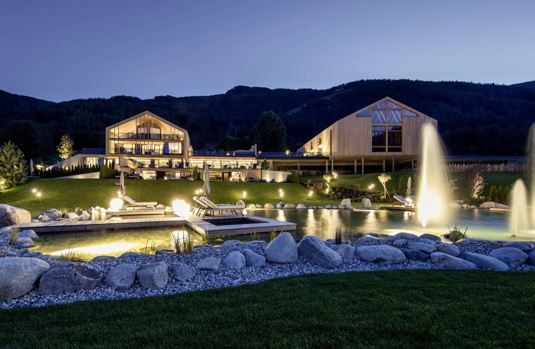 PURMONTES Private Luxury Chalet bei Nacht mit beleuchtetem Garten, Springbrunnen und Bergkulisse im Sommer