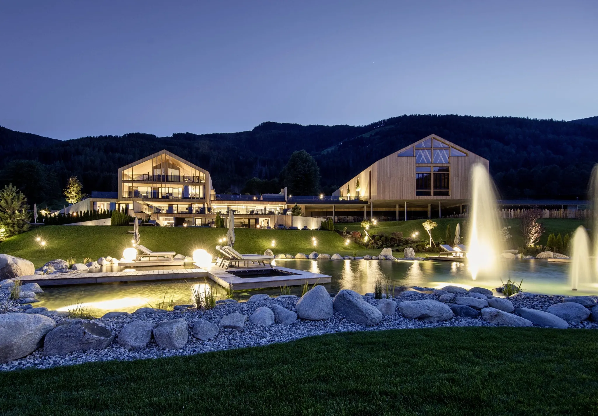 PURMONTES Private Luxury Chalet bei Nacht mit beleuchtetem Garten, Springbrunnen und Bergkulisse im Sommer
