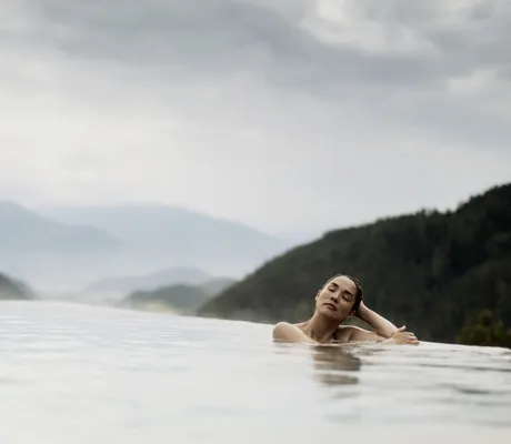 Frau entspannt im Outdoor-Infinitypool des PURMONTES Private Luxury Chalet mit Blick auf bewaldete Berge und nebligen Himmel