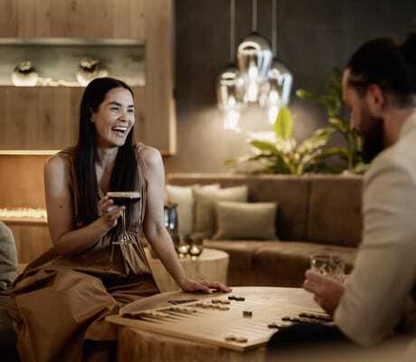 Paar sitzt an einem Holztisch im Lounge-Bereich des PURMONTES Private Luxury Chalet und spielt ein Brettspiel, Frau hält ein Getränk in der Hand