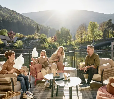 Familie sitzt auf Terrasse des Falkensteiner Family Resort Lido, umgeben von Bergen und Wald und genießt Zuckerwatte und Getränke