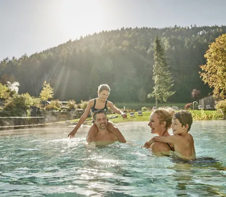 Familie mit zwei Erwachsenen und zwei Kindern spielt im Außenpool des Falkensteiner Family Resort Lido vor bewaldetem Hügel im Sonnenlicht