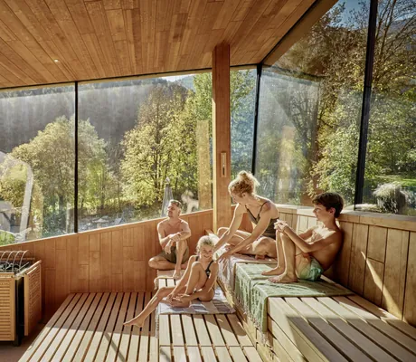 Familie mit zwei Kindern entspannt in der Sauna des Falkensteiner Family Resort Lido mit großen Fenstern und Blick auf grüne Bäume und Wiesen