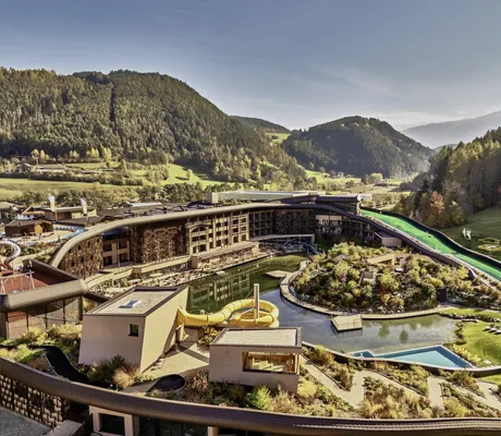 Luftaufnahme des Falkensteiner Family Resort Lido mit Außenpool und Wasserrutsche, umgeben von bewaldeten Bergen