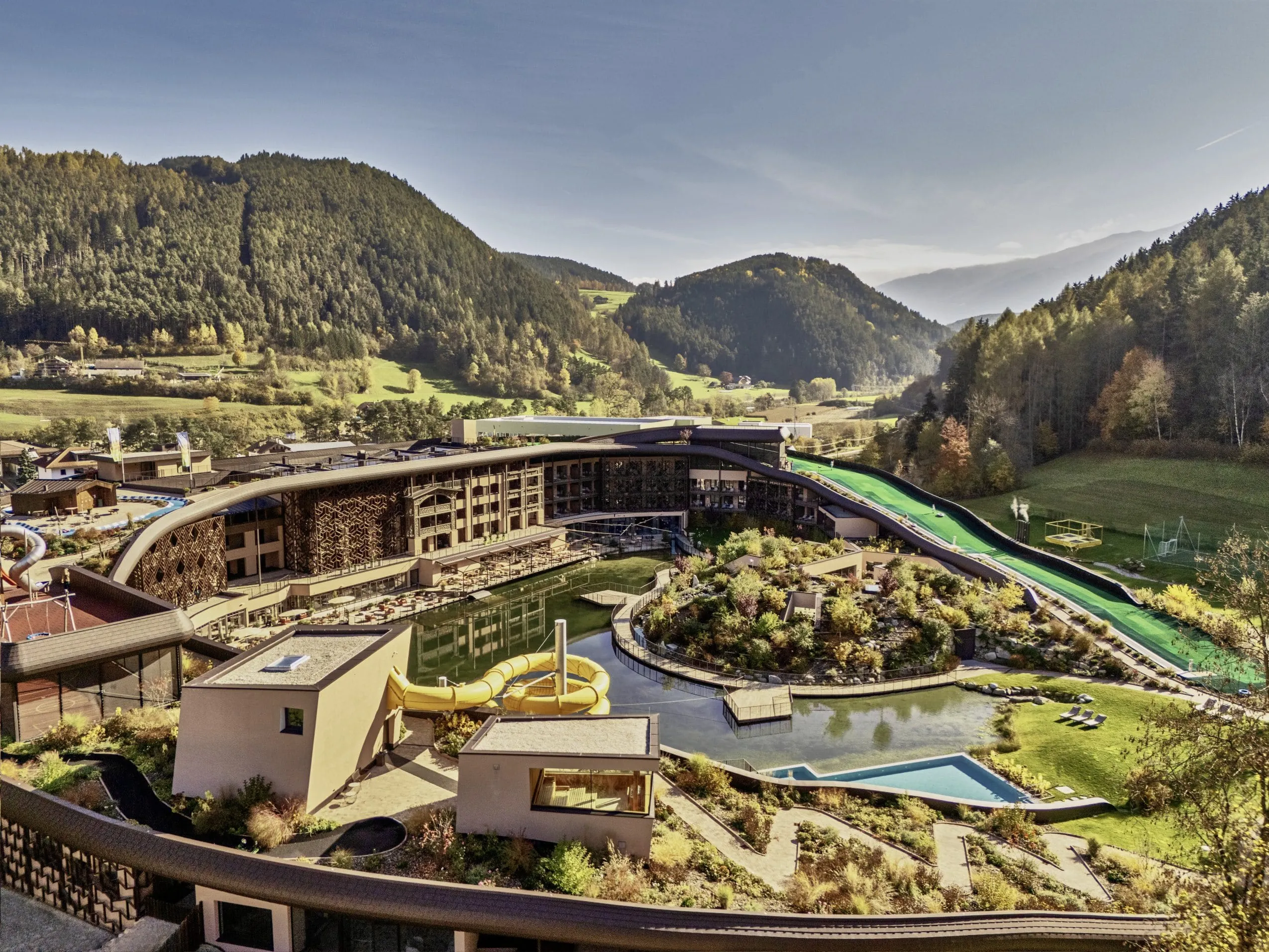 Luftaufnahme des Falkensteiner Family Resort Lido mit Außenpool und Wasserrutsche, umgeben von bewaldeten Bergen