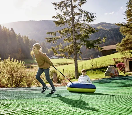 Mädchen zieht Jungen auf einem aufblasbaren Rutschreifen auf einer grünen Kunststoffmatte im Familienhotel Falkensteiner Family Resort Lido