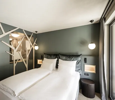 Modernes Hotelzimmer mit Doppelbett, khakifarbenen Wänden, Baumstruktur-Dekor und runden Hängelampen im Falkensteiner Family Resort Lido