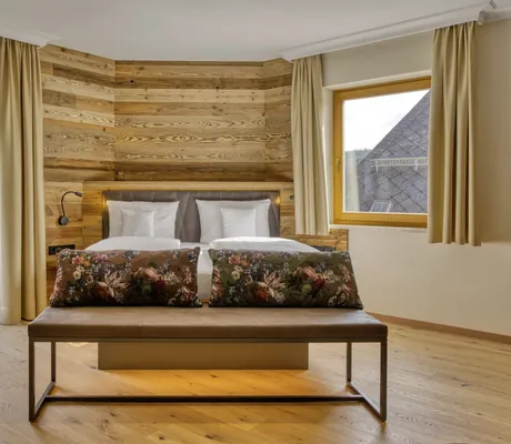 Doppelbett mit weißen Kissen in einem Zimmer des Wellnesshotel Hofbräuhaus mit Holzverkleidung und zwei Fenstern mit Blick auf bewaldete Berge und Dorf