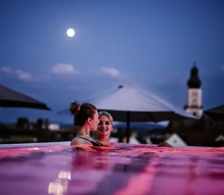 Frauen entspannen im beleuchteten Outdoorpool des Wellnesshotels Randsbergerhof bei Abendstimmung mit Vollmond und Kirchturm im Hintergrund