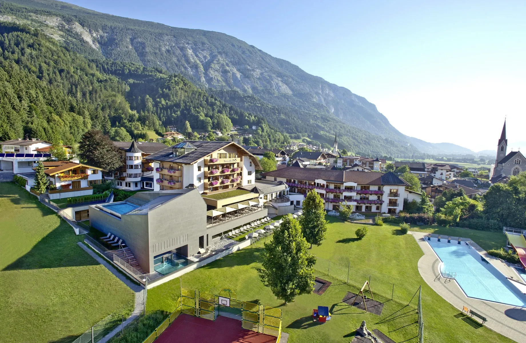 Außenansicht des Hotel Schwarzbrunn mit Außenpool, Spielplatz und Basketballfeld vor bewaldetem Bergmassiv im Sommer