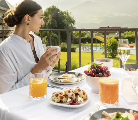 Frau in weißem Hemd sitzt auf Balkon des Hotel Schwarzbrunn mit Bergblick, Tasse in der Hand, Frühstückstisch mit Spiegeleiern, Waffeln, Trauben, Orangensaft und Sektglas
