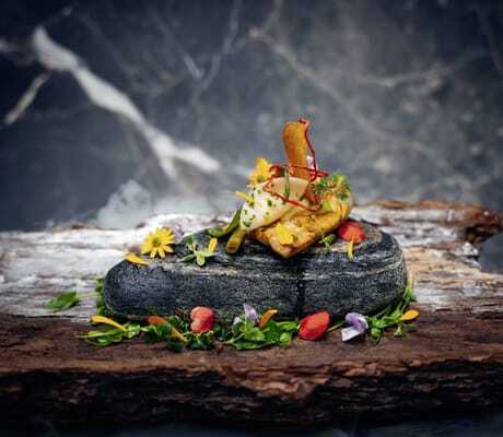 Gourmetgericht des Trofana Royal Resort mit Blüten und Kräutern auf schwarzem Stein und Holzplatte angerichtet