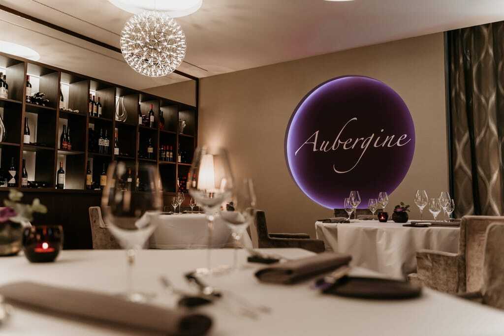 Restaurant Aubergine: Weingläser auf Tischen mit weißen Tischtüchern, im Hintergrund ein Weinregal