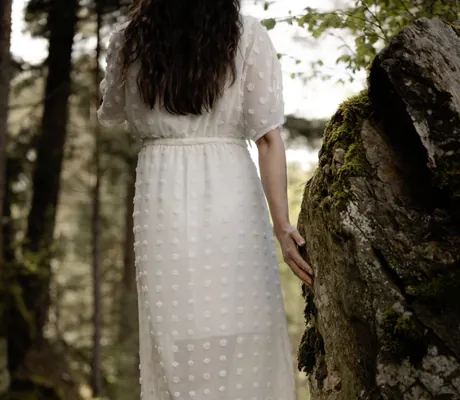 Frau mit langen dunklen Haaren in langem, weißem Kleid mit Tupfenmuster, barfuß im Wald, Hand an moosbedecktem Felsen in der Nähe des Chalet Resort ZU KIRCHWIES