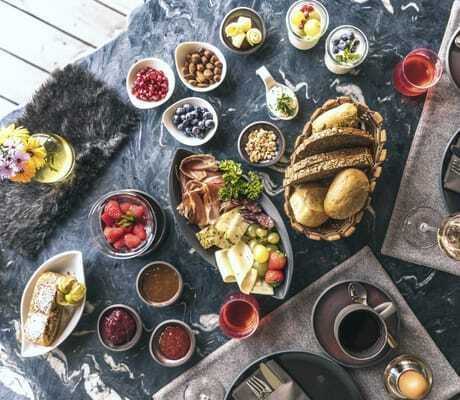 Frühstückstisch mit Brot, Schinken, Käse, Beeren, Nüssen, Marmeladen und zwei gedeckten Plätzen mit Kaffeetassen und Eierbechern im Chalet Resort ZU KIRCHWIES