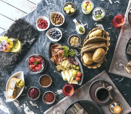 Frühstückstisch mit Brot, Schinken, Käse, Beeren, Nüssen, Marmeladen und zwei gedeckten Plätzen mit Kaffeetassen und Eierbechern im Chalet Resort ZU KIRCHWIES