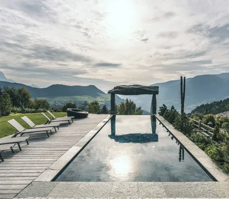 Outdoor-Infinitypool mit Liegen auf der linken Seite, umgeben von Bergen und bewölktem Himmel im Chalet Resort ZU KIRCHWIES