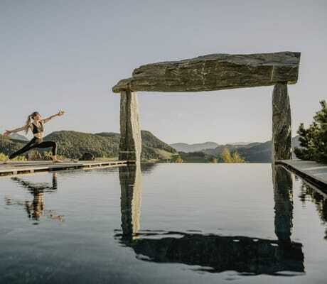 Frau in Sportkleidung macht Yoga-Pose am Rand des Infinitypools des Chalet Resort ZU KIRCHWIES mit Berglandschaft und Steinbogen im Hintergrund