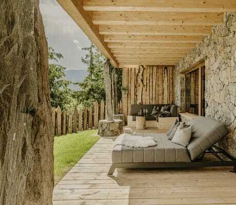 Terrasse eines Chalets im Chalet Resort ZU KIRCHWIES mit Steinmauer, Holzboden, grauer Relaxliege mit Kissen und Fell, Sofa und Holztisch, Blick auf Bäume und Berge