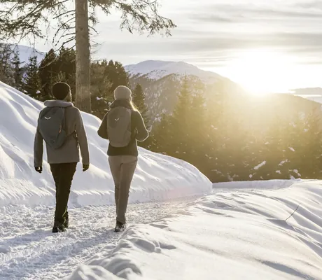 Zwei Personen mit Rucksäcken wandern auf einem verschneiten Pfad in den Bergen bei Sonnenuntergang in der Nähe des Chalet Resort ZU KIRCHWIES