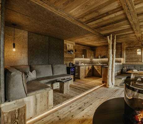Wohn- und Essbereich mit Holzwänden und -boden, grauem Ecksofa, rustikalem Holztisch, Kamin mit Glasumrandung und integrierter Küchenzeile im Chalet Resort ZU KIRCHWIES