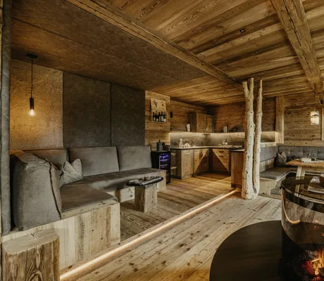 Wohn- und Essbereich mit Holzwänden und -boden, grauem Ecksofa, rustikalem Holztisch, Kamin mit Glasumrandung und integrierter Küchenzeile im Chalet Resort ZU KIRCHWIES