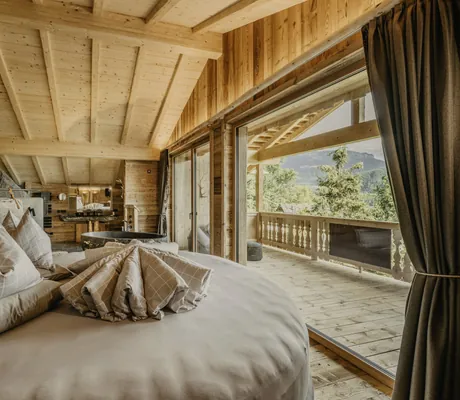 Runder Sitzbereich mit Kissen in einem Holzchalet des Chalet Resort ZU KIRCHWIES mit sichtbaren Balken, großen Fenstern und Balkon mit Wald-/Bergblick