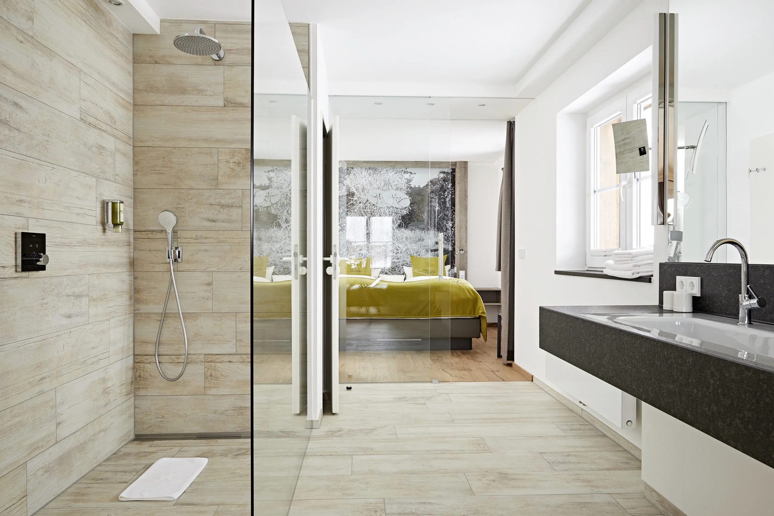 Blick durch ein Badezimmer mit Dusche und Waschtisch auf ein gro&szlig;es Doppelbett mit gelber Tagesdecke und Kissen im Gesundheitshotel Haubers Naturresort