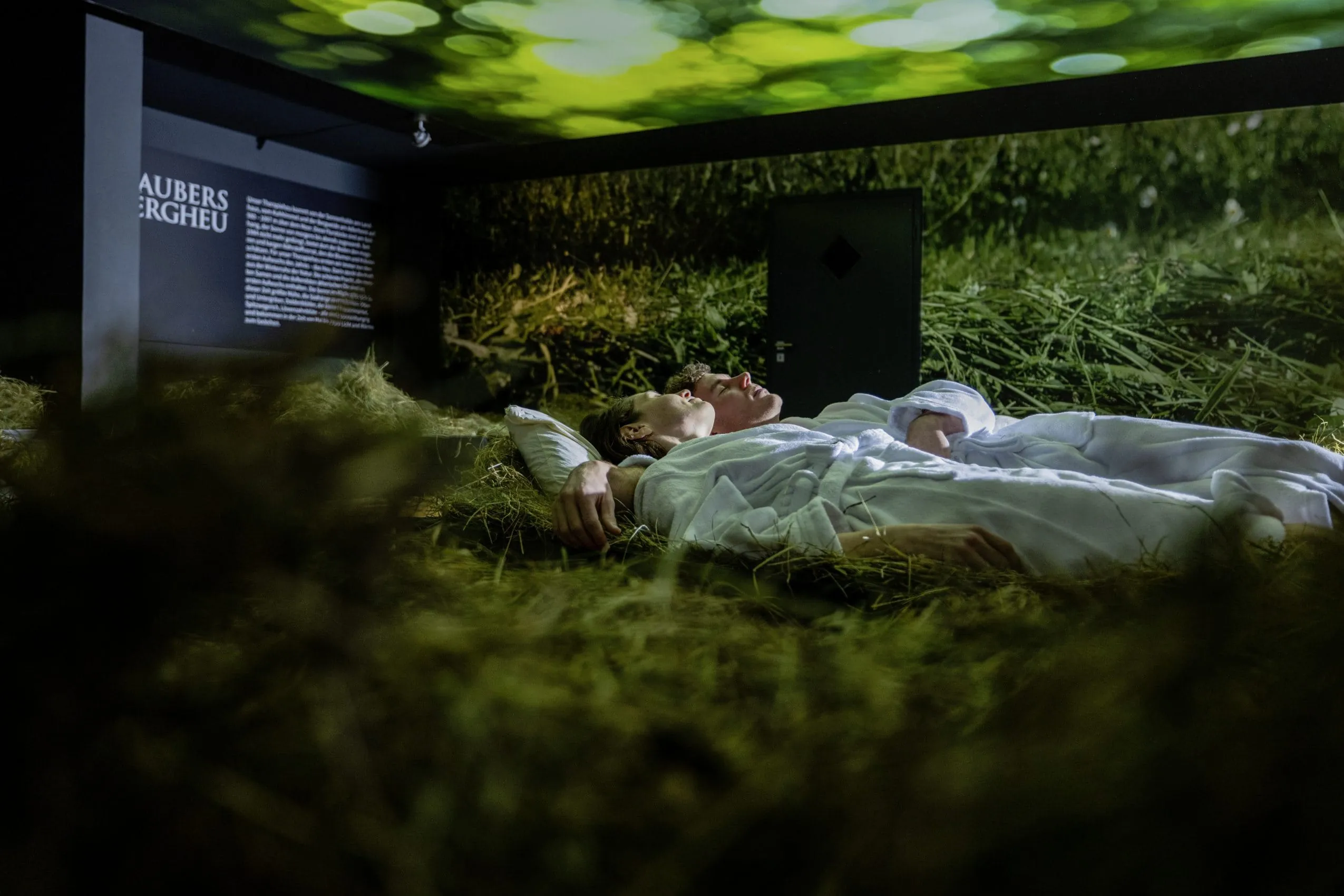Junges Paar liegt in wei&szlig;en Badem&auml;nteln im Heubett des Haubers Naturresort, an W&auml;nden und Decke sind gr&uuml;ne Licht-Projektionen