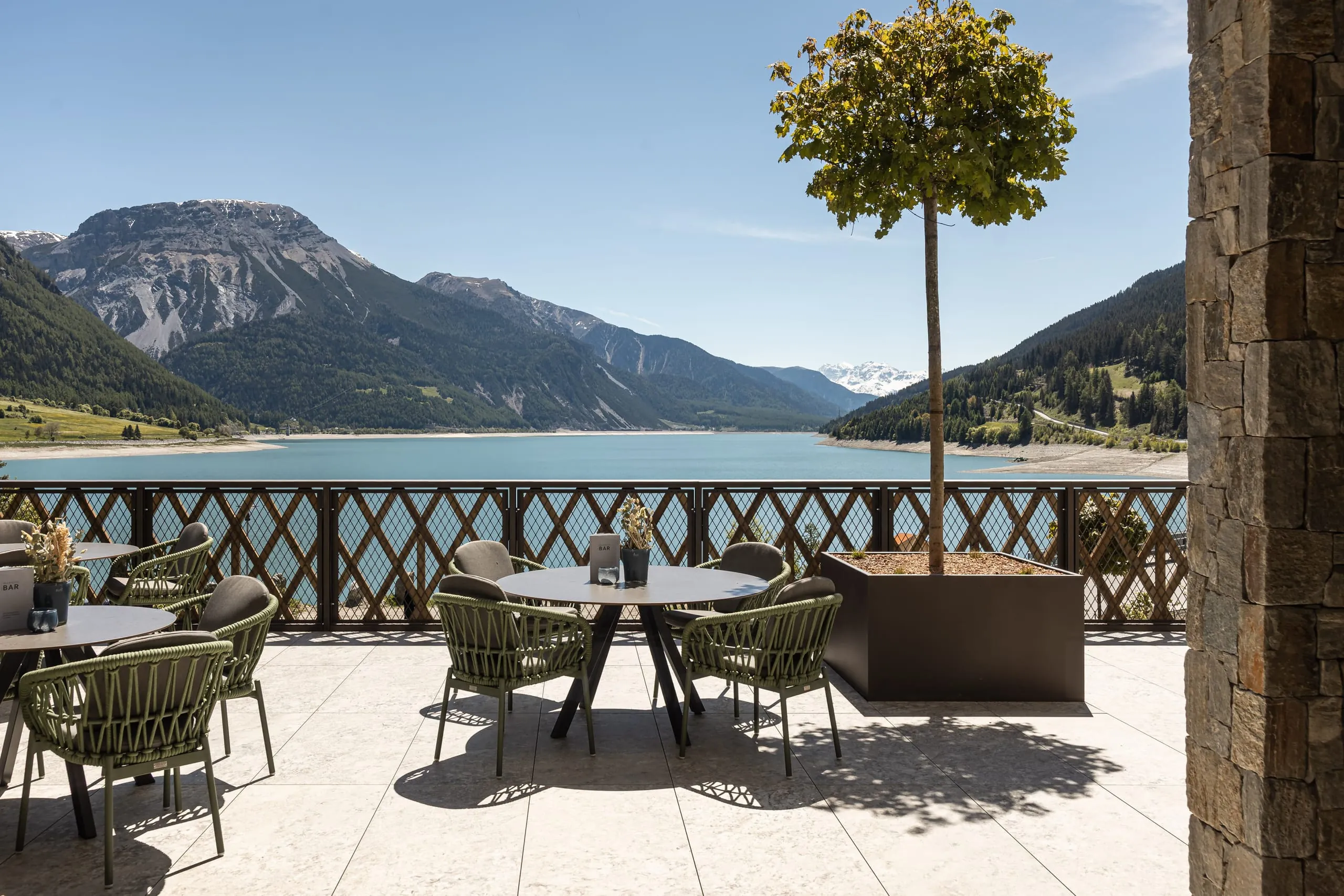 Sonnenterrasse des Gerstl Family Retreat mit Tischen und Stühlen und Blick auf den Reschensee und die Berge.