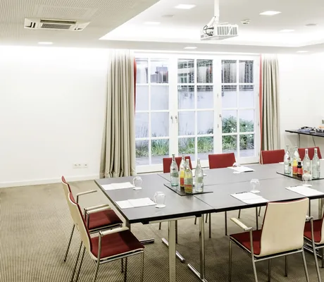 Tagungsraum im Wald & Schlosshotel Friedrichsruhe mit rechteckigem Tisch, Stühlen mit rotem Polster, Getränkeflaschen und Gläsern und großen Fenstern mit Vorhängen