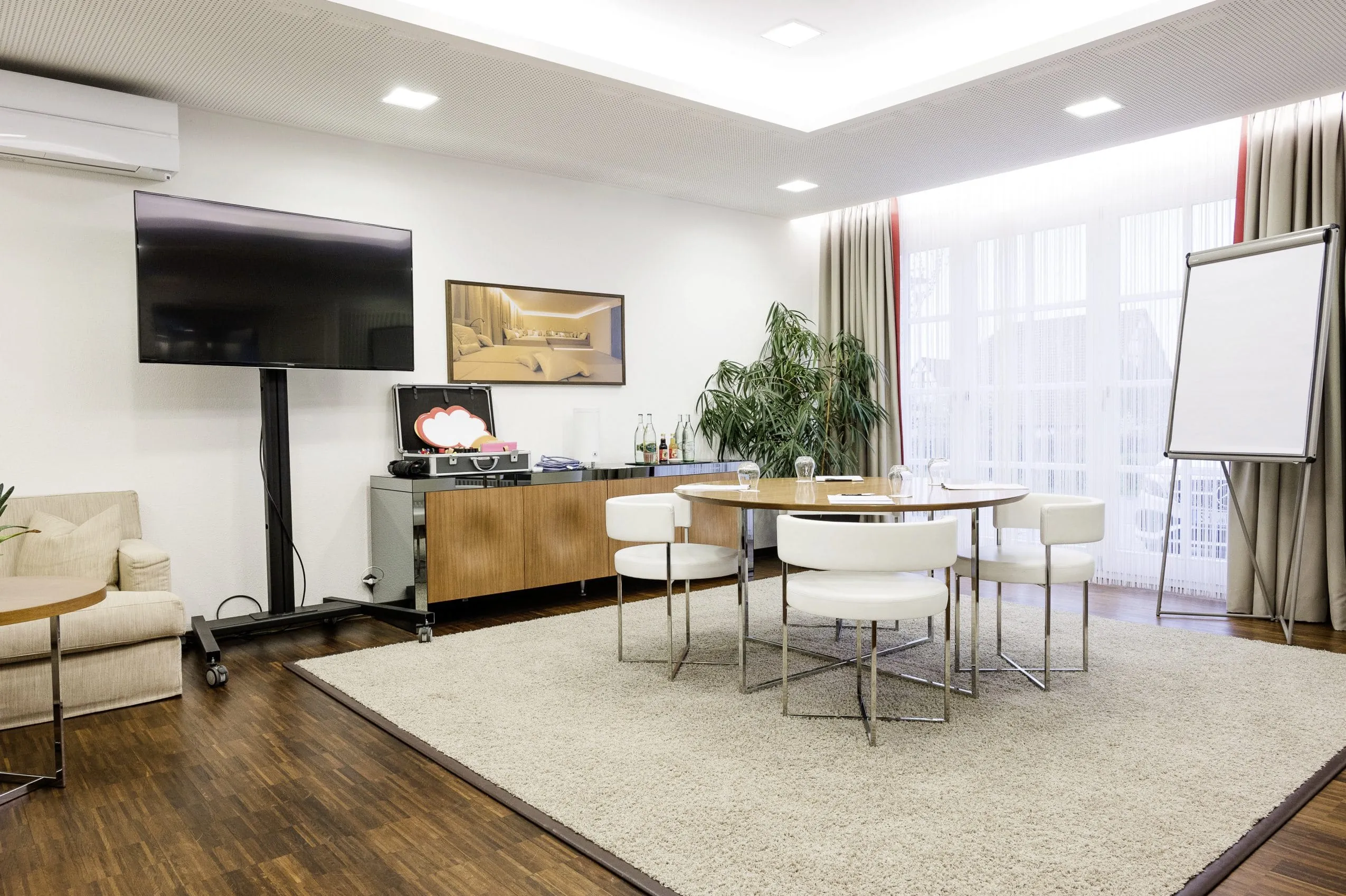 Konferenzraum mit rundem Holztisch, sechs wei&szlig;en St&uuml;hlen, gro&szlig;em Fernseher, Flipchart und Pflanze im Wald & Schlosshotel Friedrichsruhe