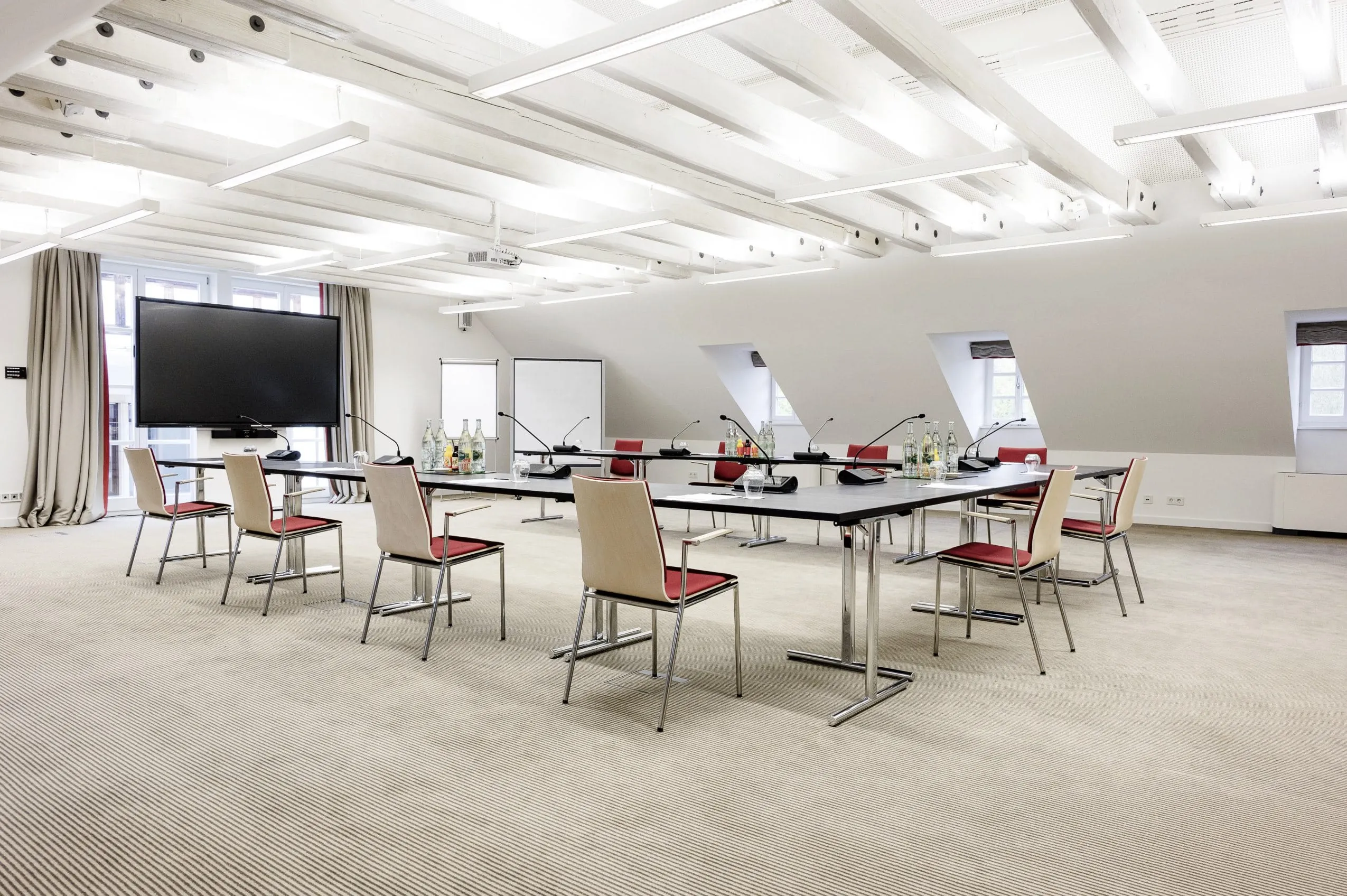 Heller Tagungsraum im Wald & Schlosshotel Friedrichsruhe mit U-f&ouml;rmiger Tischanordnung, St&uuml;hlen mit roten Sitzfl&auml;chen, gro&szlig;em Bildschirm und Flipchart