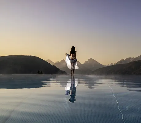 Frau mit weißem Hemd steht im Sonnenuntergang am Beckenrand des Outdoorpools im Alpen Tesitin