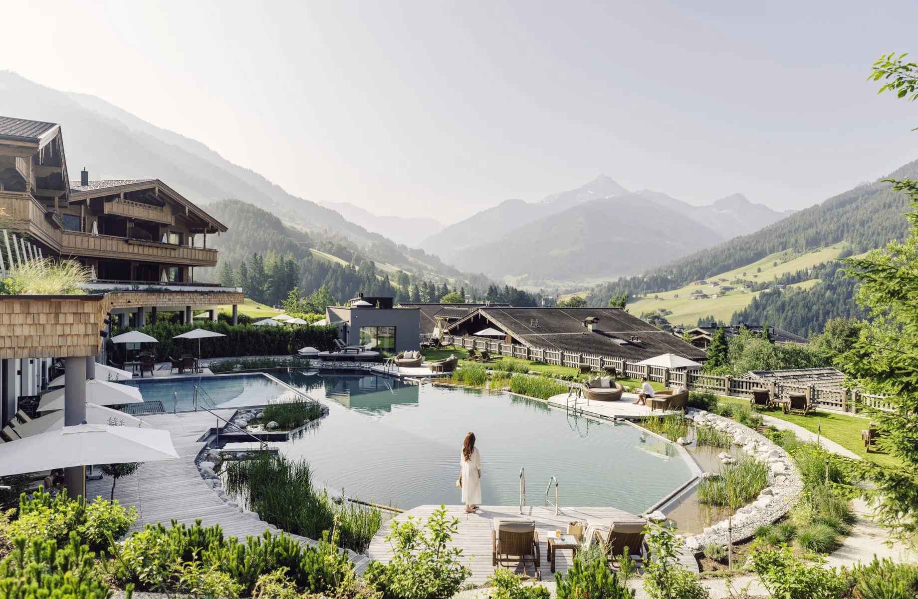 Blick auf den Naturbadesee des Der Böglerhof – pure nature spa resort und umliegende Berge, Frau steht am Rand des Sees