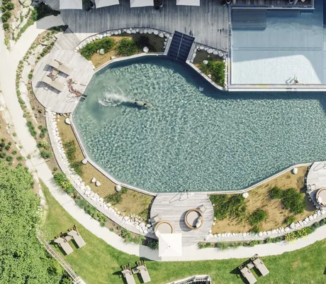 Luftaufnahme des Naturbadesees und Outdoorpools im Der Böglerhof – pure nature spa resort, umgeben von Holzdecks, Liegen und Sonnenschirmen