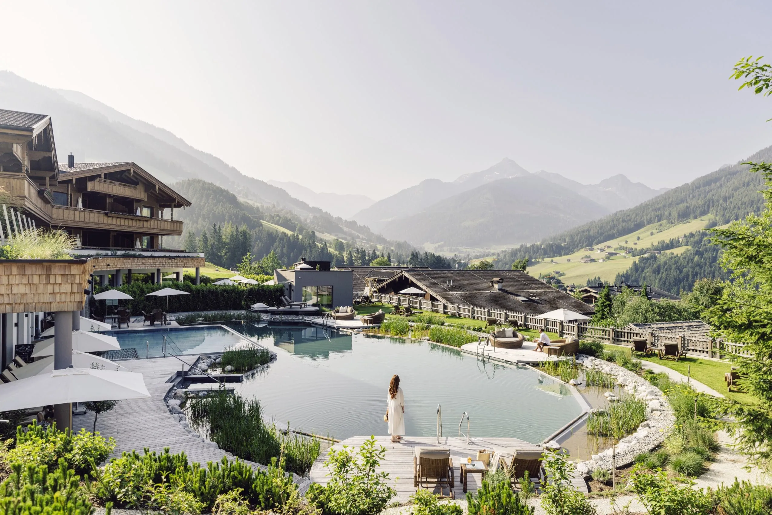 Blick auf den Naturbadesee des Der Böglerhof – pure nature spa resort und umliegende Berge, Frau steht am Rand des Sees