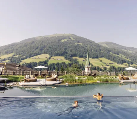 Zwei Personen schwimmen im Outdoorpool des Der Böglerhof – pure nature spa resort mit Blick auf die Berge und eine Kirche im Hintergrund