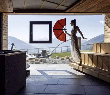 Saunameisterin mit großem roten Fächer steht am Panoramafenster der Eventsauna im Das Gerstl Alpine Retreat