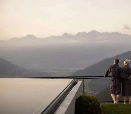 Paar in dunklen Bademänteln steht am Zaun neben dem Outdoorpool des Das Gerstl Alpine Retreat und schaut auf die Berge