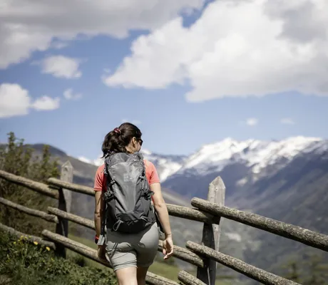 Frau mit grauem Rucksack mit Das Gerstl-Logo wandert entlang eines Holzzaunes mit Blick auf die Berge