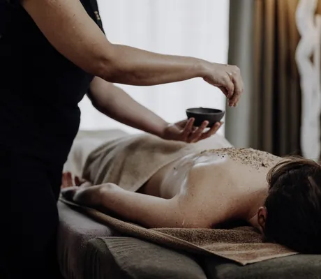 Masseurin verteilt Kräuter auf dem Rücken einer dunkelhaarigen Frau auf der Massageliege im Das.Goldberg